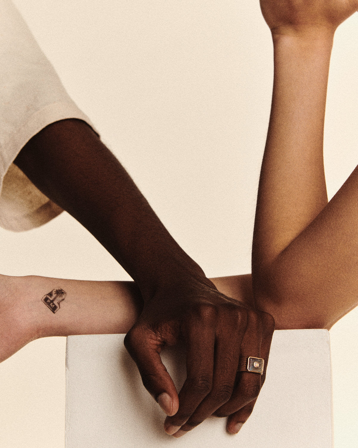 Two intertwined arms rest on a white surface. One arm, adorned with the Pascale Monvoisin ANTON N°2 MOONSTONE ring, gently grasps the other, which features a small tattoo of a house and tree against a neutral background.