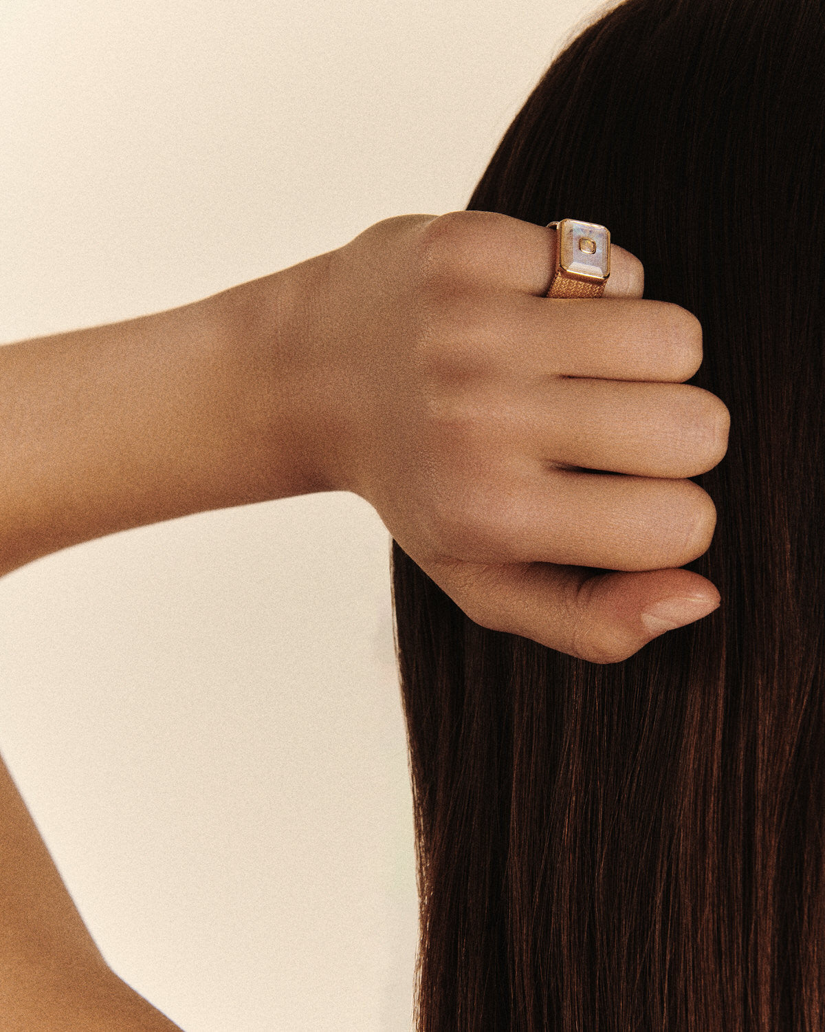 A hand with smooth skin and straight, dark brown hair features a Pascale Monvoisin ANTON N°2 MOONSTONE Ring in yellow gold. The stunning openworked ring, with its square design and captivating central pearl, gently rests against the persons head.