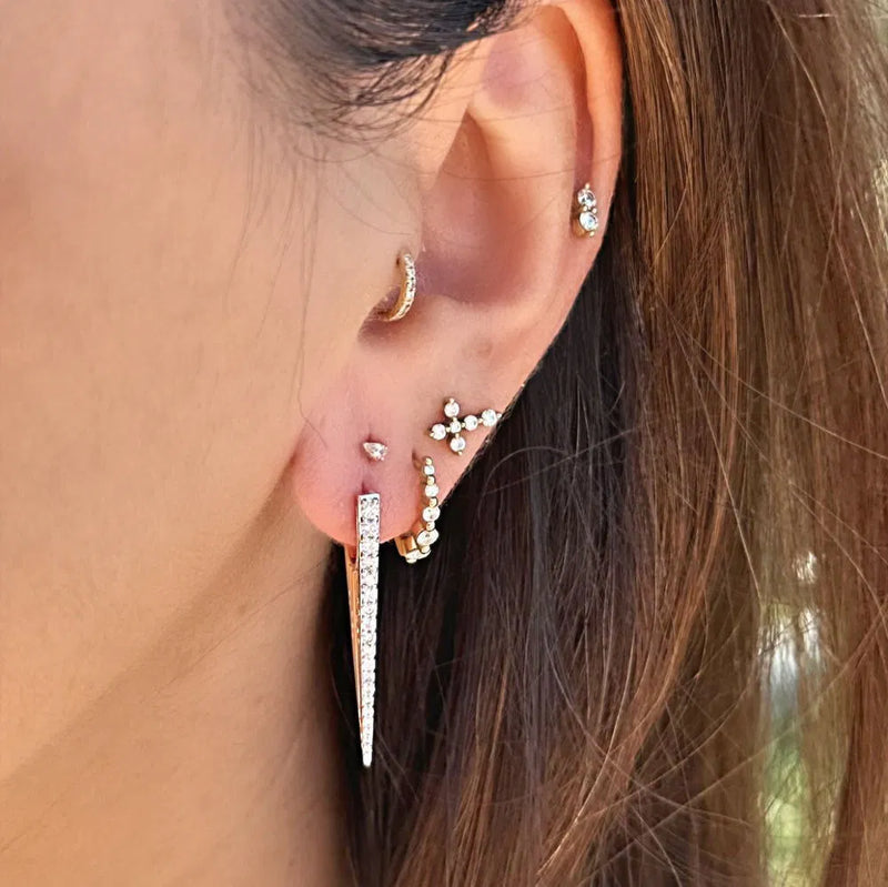 A close-up of a person’s ear with multiple piercings, featuring Three Stories Jewelry’s Elongated Diamond "V" Hoop Earrings in 14K yellow gold, alongside a crystal long earring, classic stud, pave diamond cluster, and a flower-shaped stud.