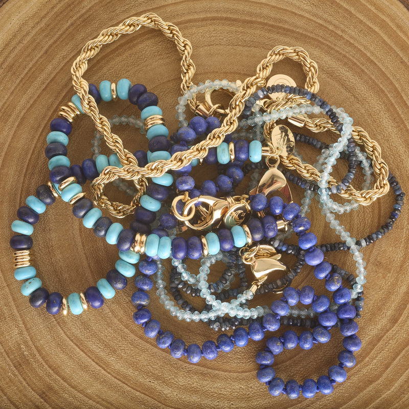The Diamond Cut Double Wrap Beaded Necklace in Kyanite by Jane Win rests on a round wooden surface, showcasing sparkling blue kyanite beads and intricate gold detailing against the wood’s visible grain.