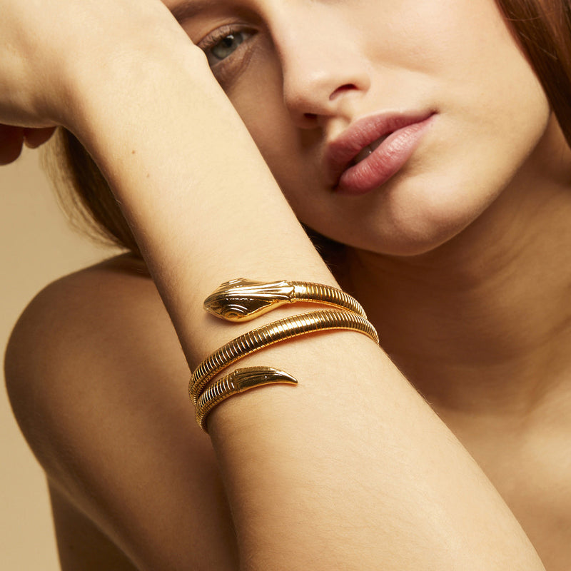 A woman rests her face on her arm, displaying the gas bijoux Serpent bracelet—a flexible gold snake design wrapped elegantly around her wrist against a soft beige background, with only her lips and part of her face visible.