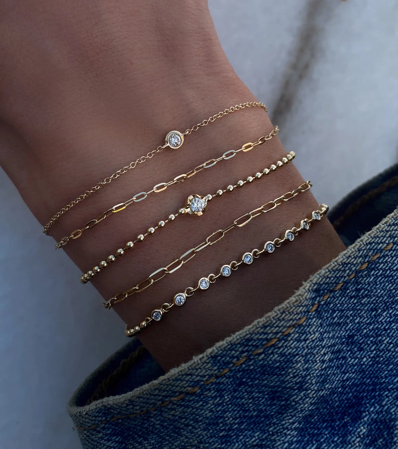 A wrist wearing five gold bracelets, including Luna Skyes 14kt gold two row diamond bezel chain bracelet, shines beneath a blue denim jacket.