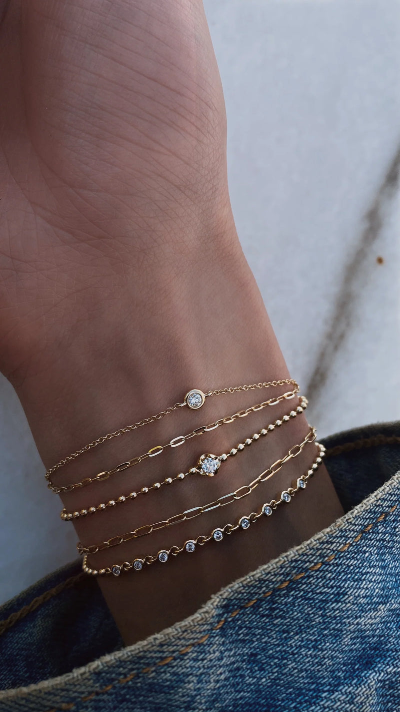 A close-up of a wrist styled with five delicate gold bracelets, featuring Luna Skyes 14kt gold two row diamond bezel chain bracelet, above a denim cuff on light marble.