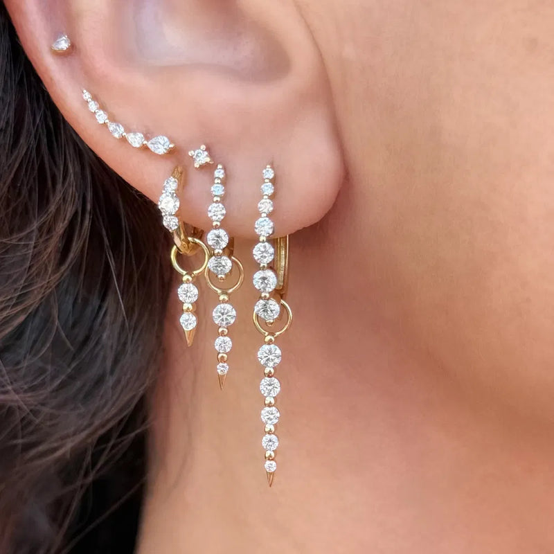 A close-up of an ear features vintage-inspired 14K yellow gold earrings from Three Stories Jewelry, adorned with Medium Sari Dagger Charms cascading down the earlobe and auricle, their delicate diamonds sparkling softly against light skin.