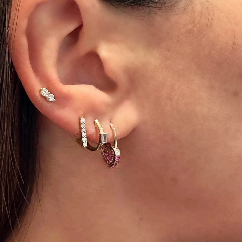 An ear adorned with multiple piercings displays a small triple stud on the upper lobe, a row of diamante studs along the edge, and eye-catching hoops. Among them, a Tiny Pave Rectangular Hoop from Three Stories Jewelry glimmers alongside diamond earrings, all set in 14K yellow gold.