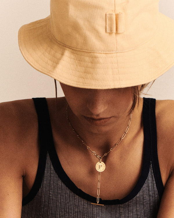 A person wearing a beige bucket hat and gray tank top with black trim looks down. They wear a Pascale Monvoisin Debbie No. 2 Necklace, personalized in 9K yellow gold with a circular pendant. Soft lighting casts gentle shadows, highlighting their exquisite accessory.