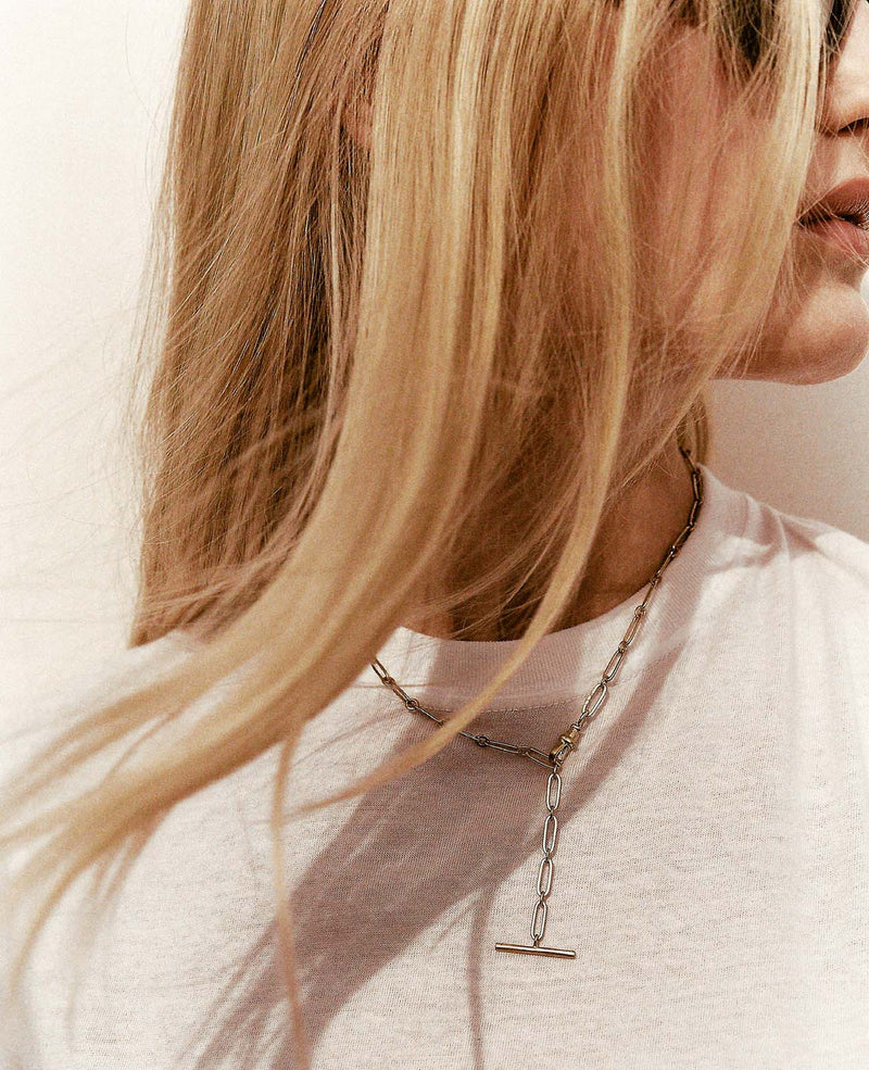 A person with long blonde hair wears a white shirt and a Debbie No. 2 Necklace by Pascale Monvoisin, featuring a 9K yellow gold bar pendant. Their hair partially covers their face as they look away from the camera.