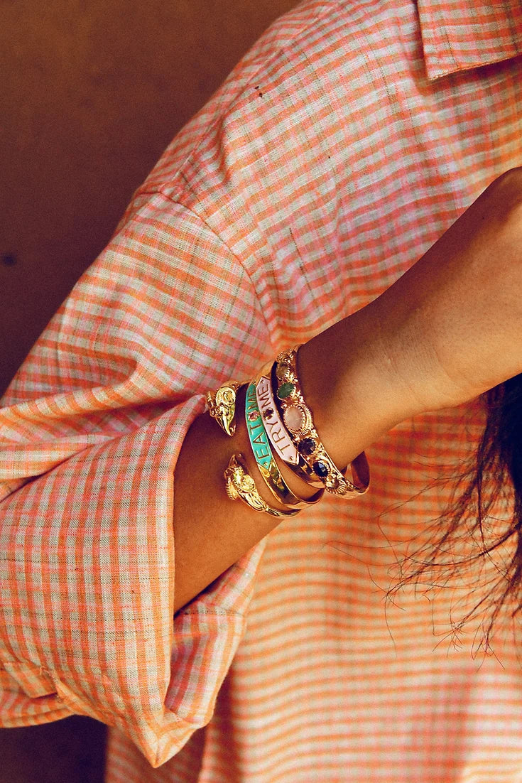 A person wearing an orange and white plaid shirt displays their forearm adorned with multiple bangles, including a standout rhodium-plated brass SYLVIE bracelet from LAltelier Plume. The slightly rolled-up sleeve enhances its reflective charm.