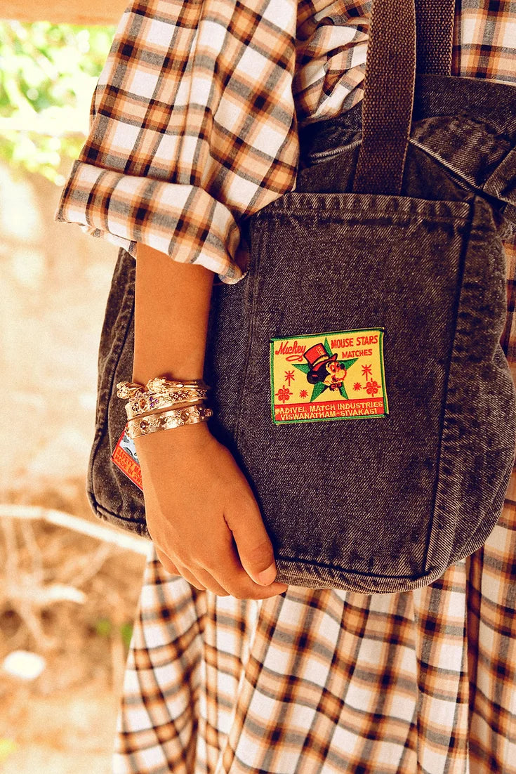 A person in a plaid dress holds a denim bag with a colorful patch of a mouse, stars, and matches. They wear several SYLVIE bracelets by LAltelier Plume, reflecting their unique style against a softly blurred background of earthy tones and greenery.