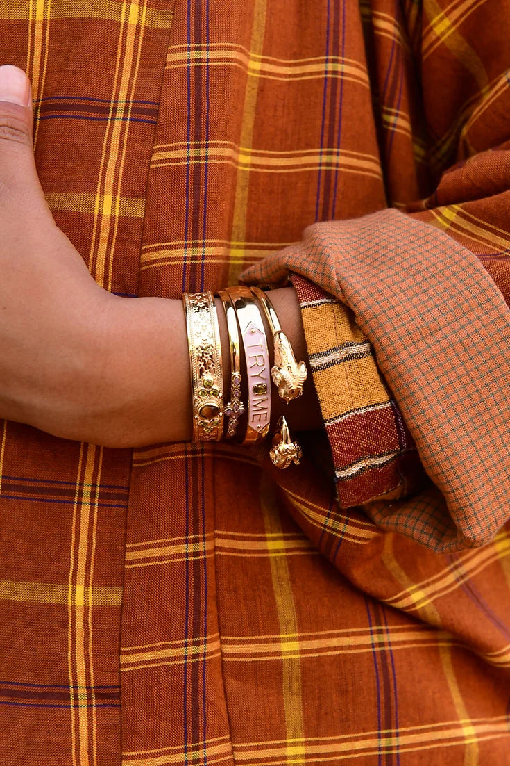 A person in a brown and orange plaid garment accessorizes with LAltelier Plumes SYLVIE bracelet, a rhodium-plated brass piece. The bracelet features the word CROWN and its reflective shine catches attention as their casually placed hand rests on the opposite arm.