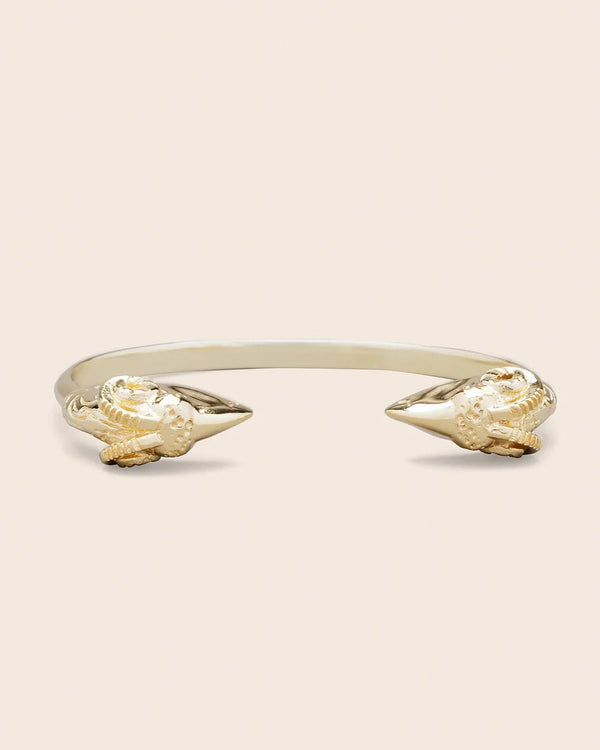 The SYLVIE bracelet by LAltelier Plume is a sleek, gold-tone cuff crafted from rhodium-plated brass with lion head designs at each end. Set against a plain beige background, it highlights intricate carvings and outstanding corrosion resistance.