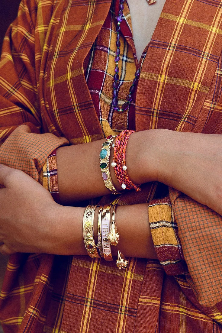 A person in a plaid orange shirt with rolled-up sleeves showcases various bracelets, including LAltelier Plumes SYLVIE rhodium-plated brass bangles and multicolored pieces with turquoise stones. Their design enhances the layered bohemian look with reflective charm.