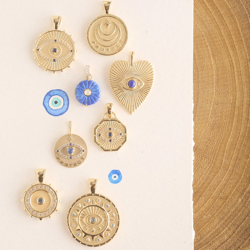 The Jane Win PROTECT Eye On Your Heart Pendants with blue evil eye designs and lapis stones are displayed on a textured white background near wood, accompanied by blue and white round evil eye charms.