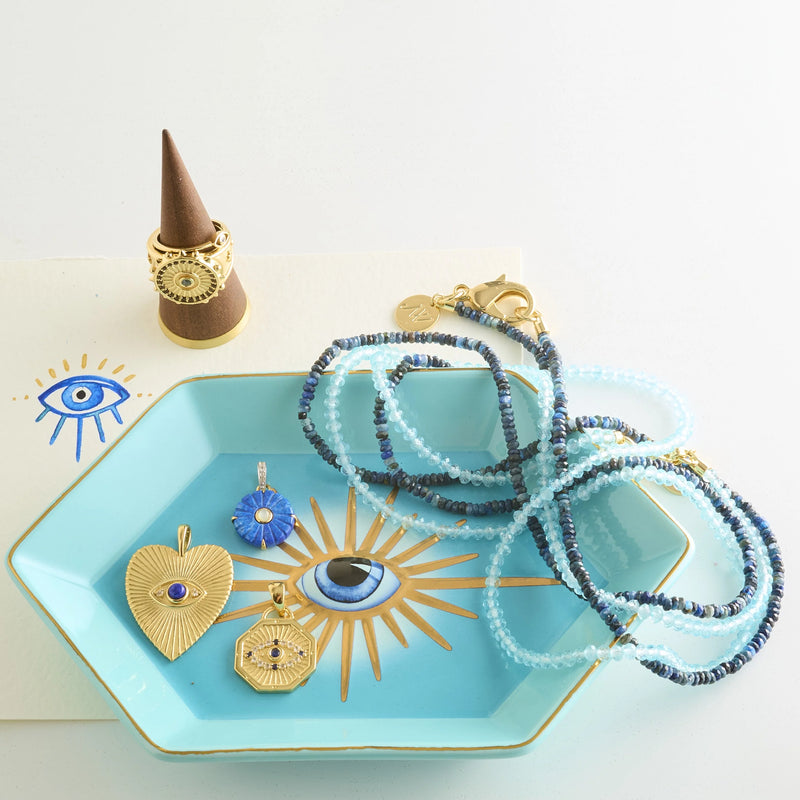 A turquoise dish with gold rays holds three gold pendants, including the Jane Win PROTECT Evil Eye Petite Embellished Pendant, a heart pendant, and blue evil eyes. Beaded blue necklaces, gold rings on a cone, and an illustrated blue eye card are nearby.