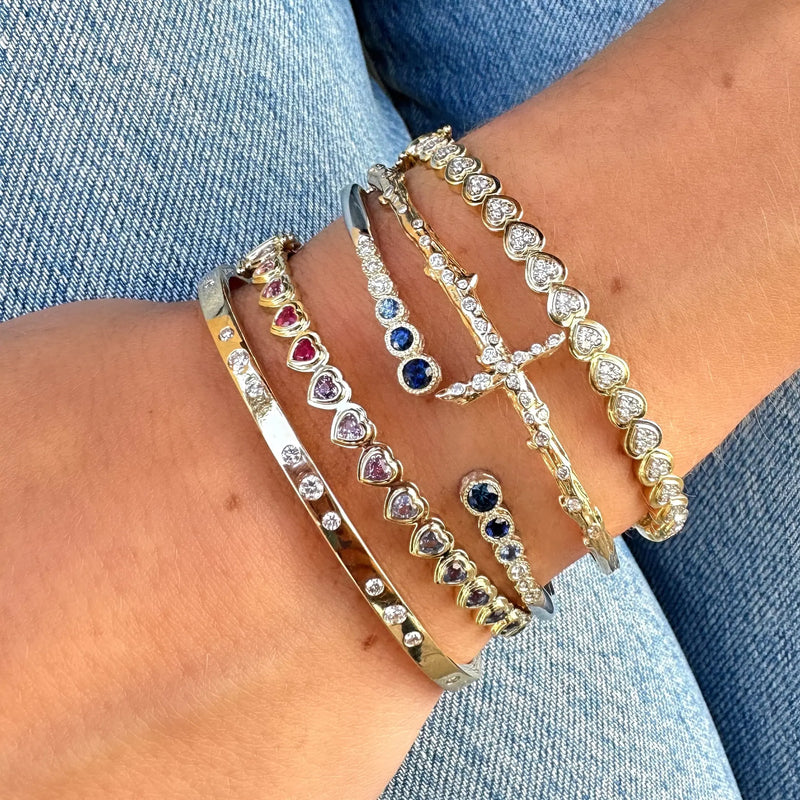 A person wears multiple gold bracelets, including the Love Explosion Sideways Pave Heart Bangle by Three Stories Jewelry, featuring round diamonds and heart-shaped stones, paired with classic blue jeans for an elegant look.