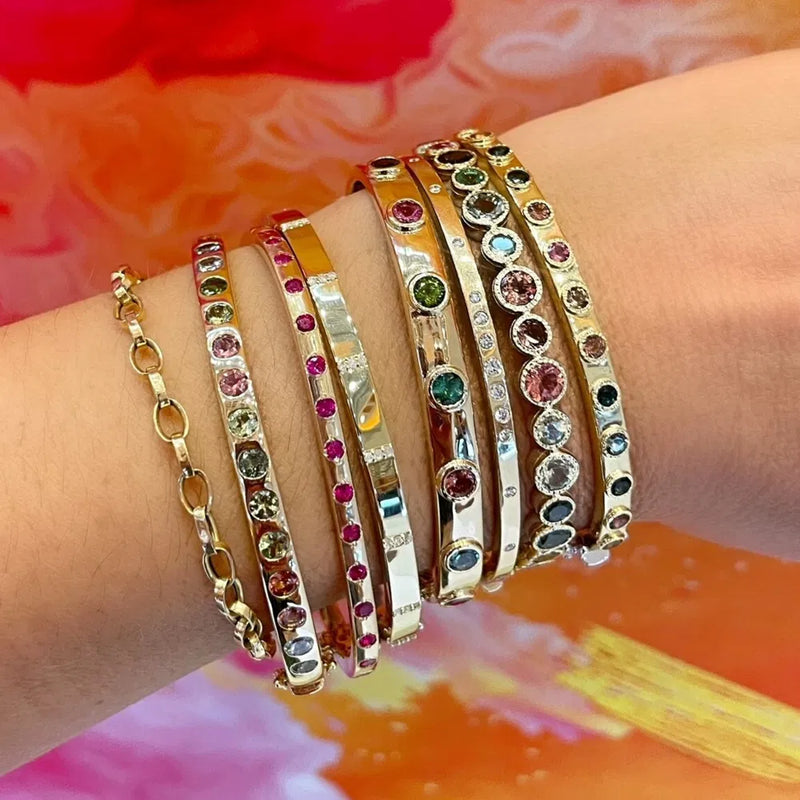 A wrist showcases the Two-Toned Flip Colored Tourmaline Bangle by Three Stories Jewelry. The piece features vibrant tourmaline in diverse settings against a vivid pink and orange backdrop.