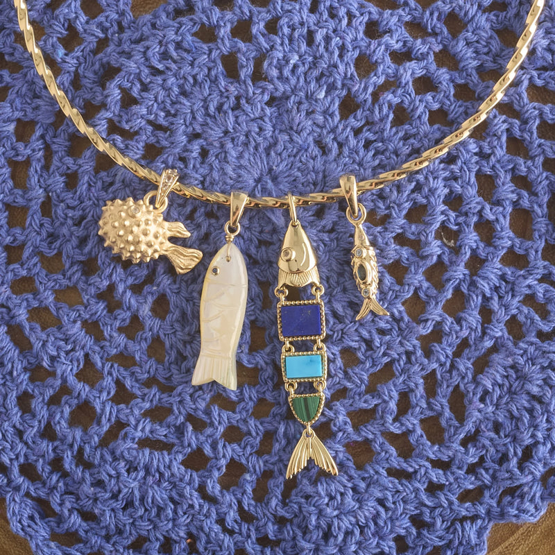 The Jane Win STRONG Carpe Diem Fish Pendant is a vintage-style gold necklace featuring four unique fish-shaped charms, beautifully displayed on a blue crocheted doily background.
