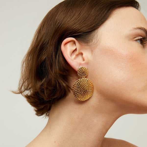 Close-up of a womans head and neck, highlighting her short brown hair and gas bijoux Onde earrings Small—handmade in France, gold-plated with two textured circular pieces, shown against a plain light background.
