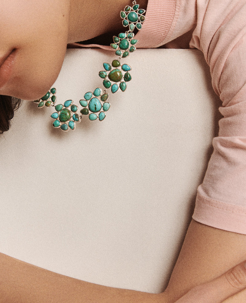 Close-up of someone wearing a light pink shirt and the Necklace GLORIA N°2 by Pascale Monvoisin, featuring green and blue floral stones with Turquoise accents on Silver 925. They rest on a white surface with their face partially out of frame.