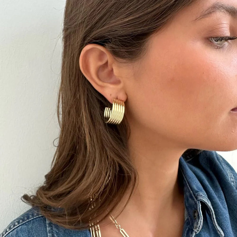 A person with long brown hair wears a denim jacket and stylish jewelry, featuring the Jennifer Zeuner brands Cairo 1 Hoops with a ribbed design, visible in a close-up side profile against a light background.