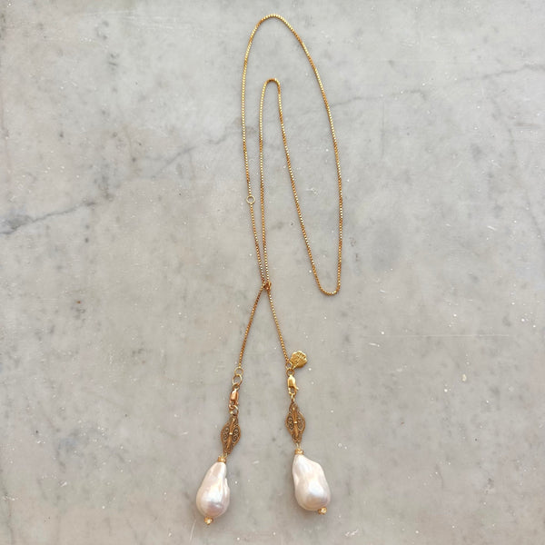 Introducing the JOHN ROSS by Bohemian Rhapsodie Paris: a graceful gold-plated lariat necklace featuring a chain with two ornamental pendants. Its intricate designs and large, irregular pearls make it an ideal meditation accessory, elegantly displayed on a light-colored marble surface.