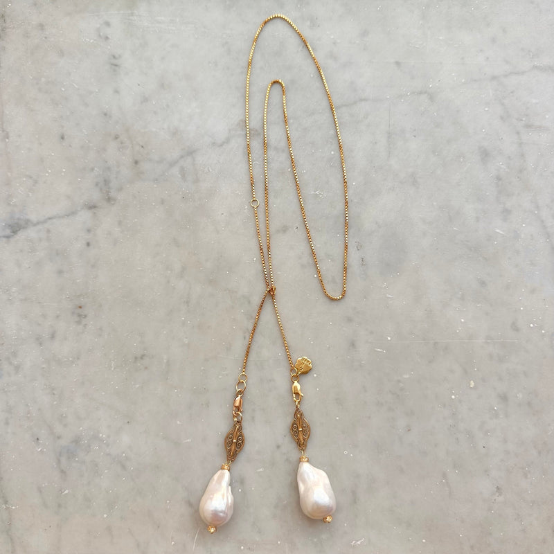 Introducing the JOHN ROSS by Bohemian Rhapsodie Paris: a graceful gold-plated lariat necklace featuring a chain with two ornamental pendants. Its intricate designs and large, irregular pearls make it an ideal meditation accessory, elegantly displayed on a light-colored marble surface.