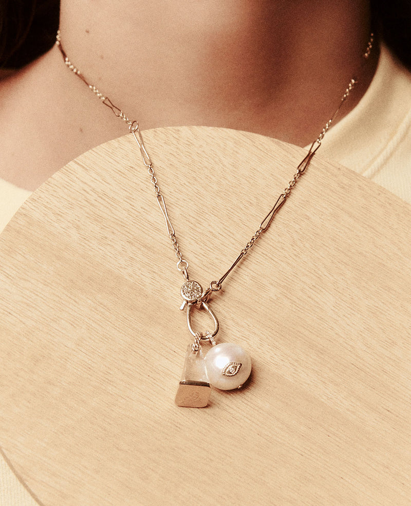 The Marcia Necklace by Pascale Monvoisin is a delicate yellow gold piece with an adjustable chain, featuring a teardrop gemstone, large pearl, and small square pendant. It elegantly rests on a wooden surface against a neutral fabric backdrop.