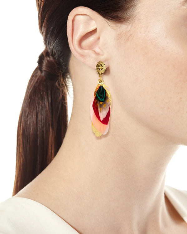 A close-up of an individual wearing the Sao Feather Drop Earrings by gas bijoux, featuring vibrant red, pink, yellow, and green feathers with elegant gold detailing. Their hair is tied back and they are dressed in a cream-colored top embellished with Swarovski crystal accents.