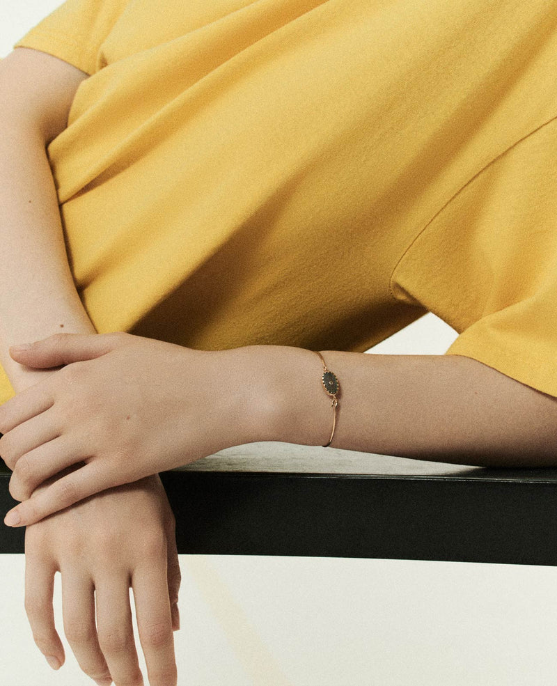 A person in a yellow shirt leans on a black table, arms crossed, showcasing the Bracelet ORSO MOONSTONE by Pascale Monvoisin, adorned with a teardrop-shaped stone. The neutral background accentuates the simplicity and elegance of the scene.