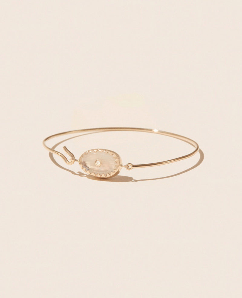 The ORSO MOONSTONE bracelet by Pascale Monvoisin showcases a slender yellow gold band adorned with an oval-shaped, translucent stone centerpiece. It elegantly rests on a neutral beige background, casting a subtle shadow.