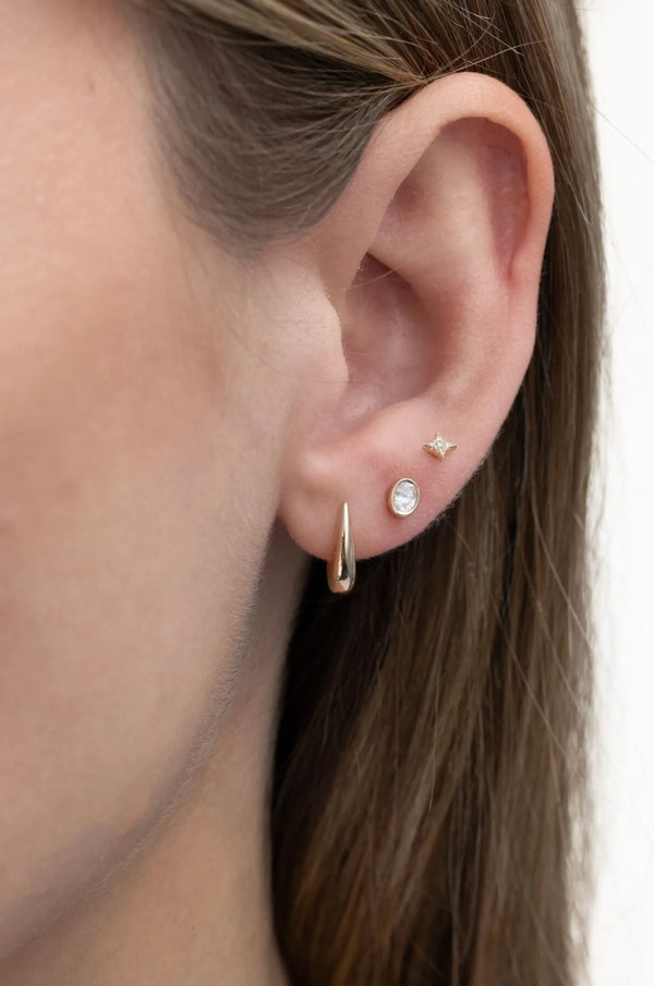 Close-up of a womans ear with three earrings: a gold spike hoop in the lobe, an ef collection Single Bezel Set Oval Diamond Stud Earring above it, and a tiny star-shaped stud higher on the ear. Brown hair frames the dainty diamond detail.