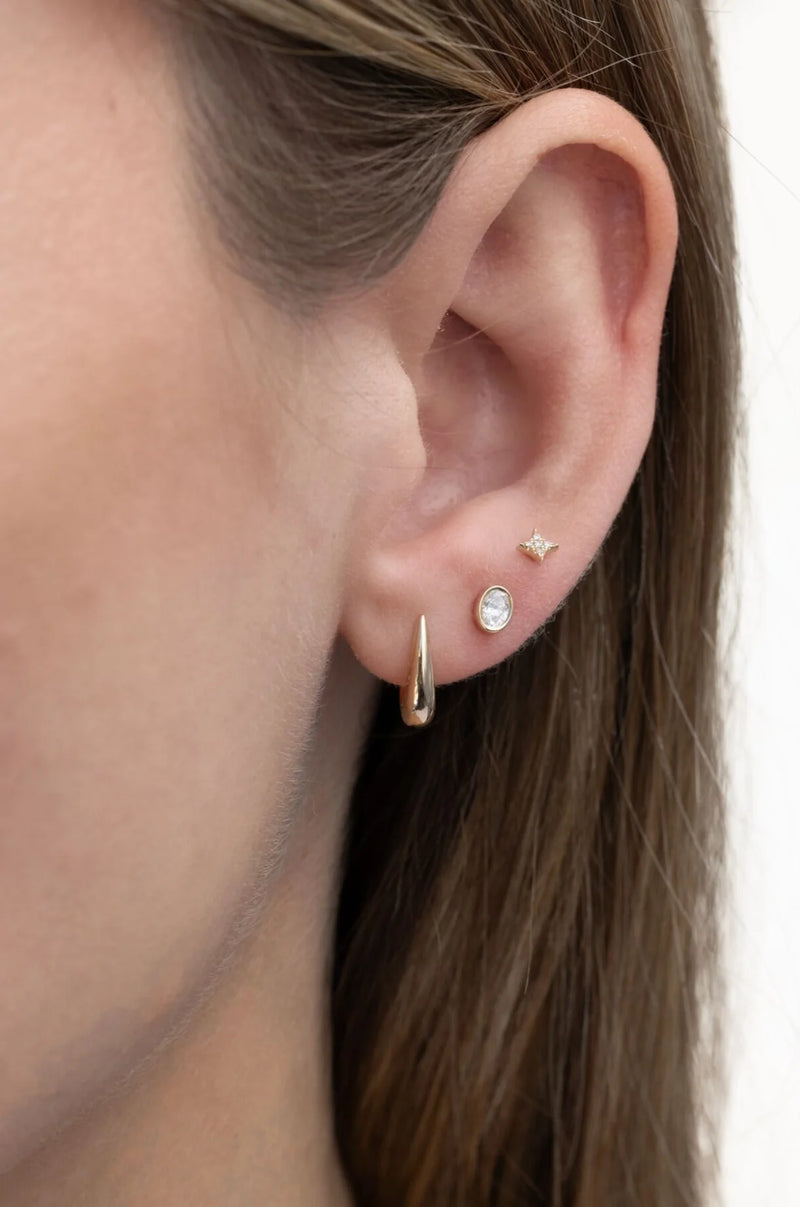 Close-up of a womans ear with three earrings: a gold spike hoop in the lobe, an ef collection Single Bezel Set Oval Diamond Stud Earring above it, and a tiny star-shaped stud higher on the ear. Brown hair frames the dainty diamond detail.