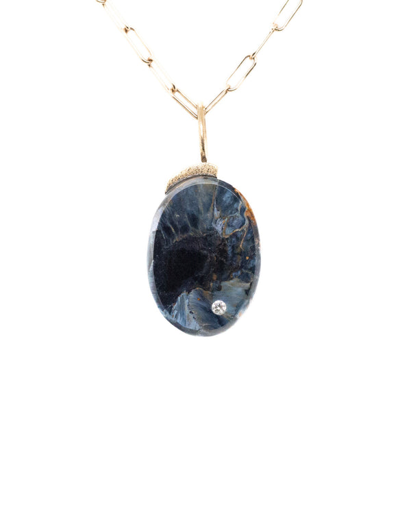 The Karen Luvaas Pietersite Oval Charm is a gold chain necklace featuring an oval, dark blue pietersite stone with swirling patterns and a small round clear gem set near the bottom.