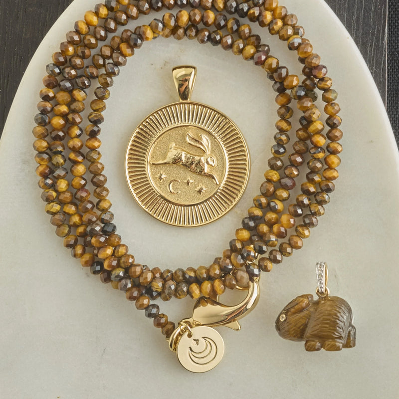 The Jane Win Double Wrap Diamond Cut Beaded Necklace in Tiger's Eye is coiled on a light oval surface, featuring a large gold rabbit pendant, a smaller gold charm, and a detailed brown rabbit charm.