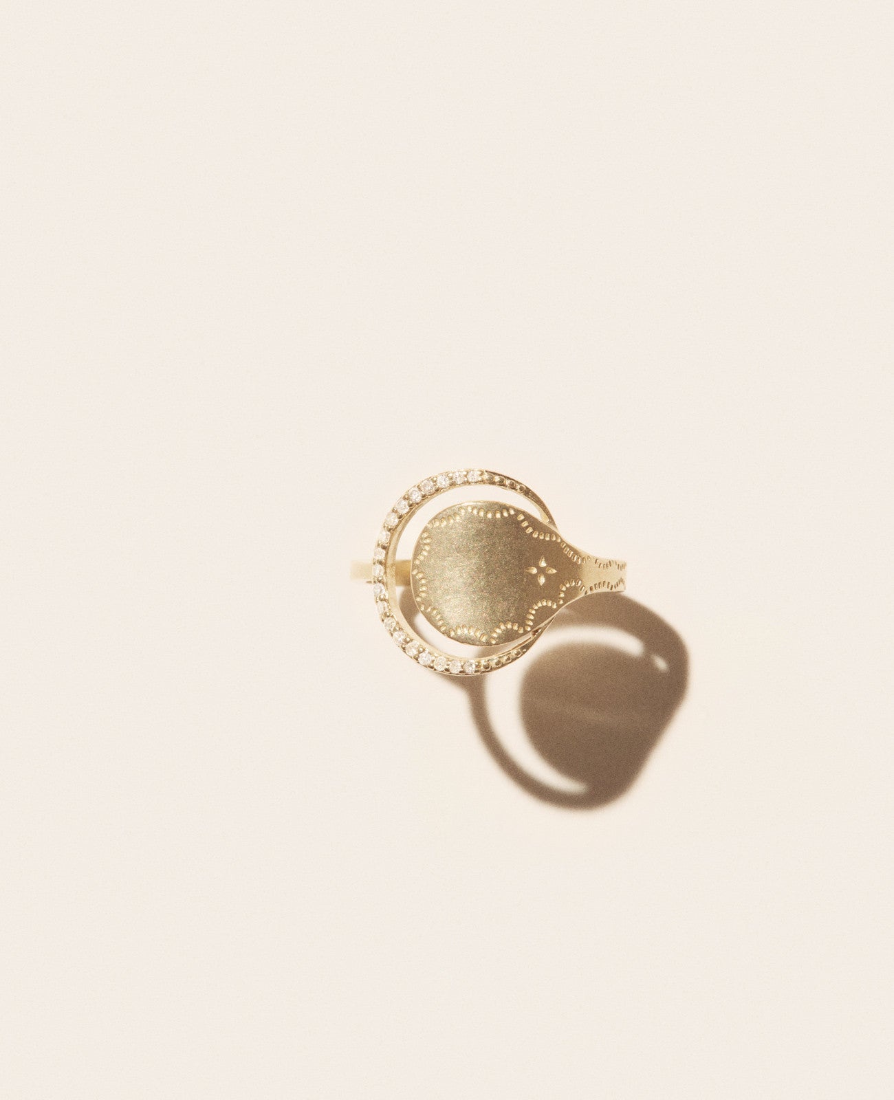 The Rio Ring by Pascale Monvoisin, featuring a textured gold oval centerpiece adorned with small clear stones and hints of turquoise, casts a soft shadow against a pale background.