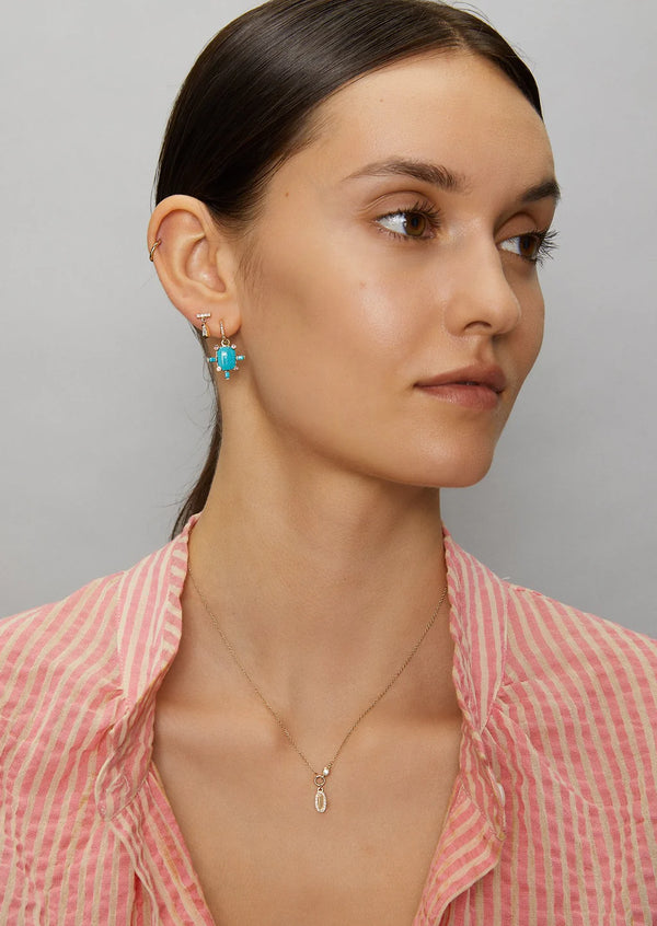 A woman with straight brown hair and natural makeup wears layered Scosha Juno hoop earrings featuring turquoise charms, a 14k gold pendant necklace, and a pink and white striped shirt. She looks to the side.