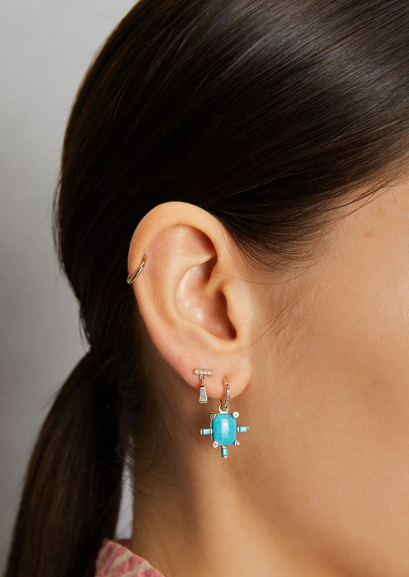 Close-up of a womans ear with three earrings: a 14k gold hoop on the upper ear, a stud, and the Scosha Juno Charm with Turquoise—a dangling earring featuring a rectangular turquoise stone and small clear stones on the lobe.