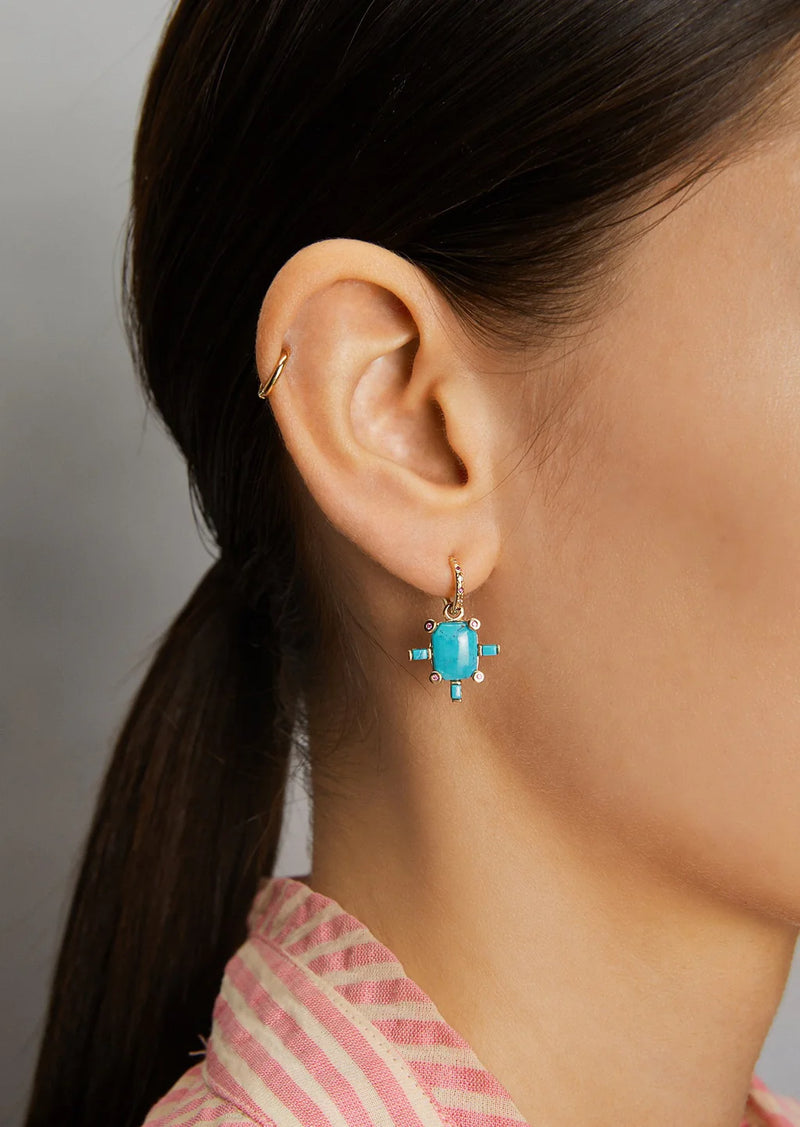 Close-up of a woman’s ear wearing the Scosha Juno Charm with Turquoise, a 14k gold earring accented by small blue gems. Her brown hair is pulled back, and she wears a pink and white striped top.