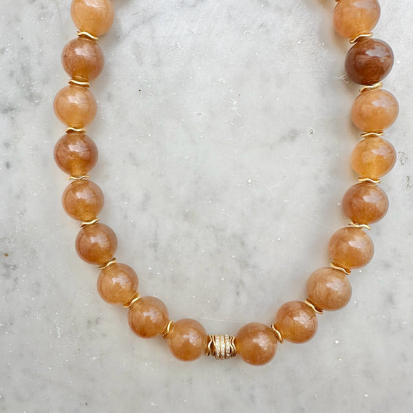 The SLY Camel necklace by Bohemian Rhapsodie Paris is displayed on a light marble surface, showcasing its round, polished amber beads linked by gold-plated rings and accented with a decorative gold clasp at the center.