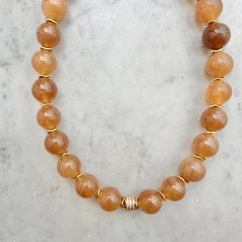 The SLY Camel necklace by Bohemian Rhapsodie Paris is displayed on a light marble surface, showcasing its round, polished amber beads linked by gold-plated rings and accented with a decorative gold clasp at the center.