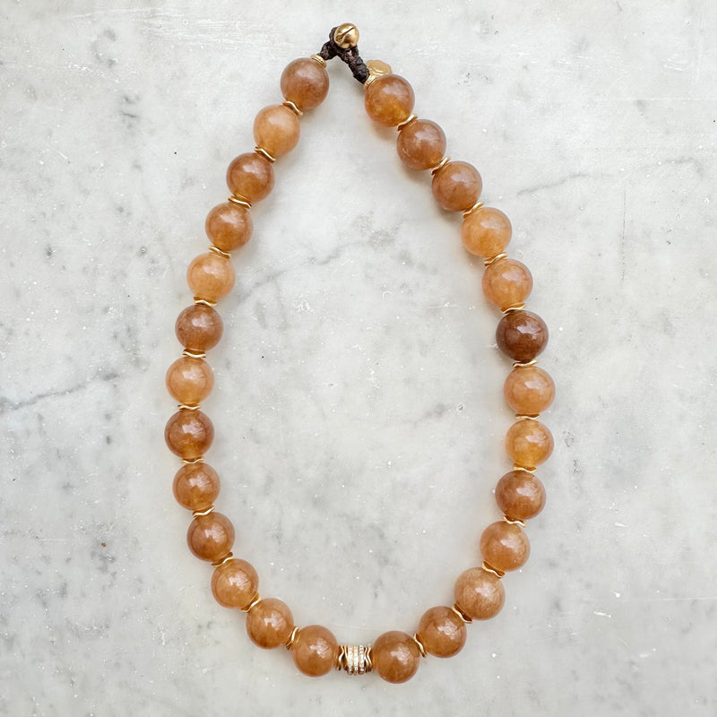 The SLY Camel necklace from Bohemian Rhapsodie Paris features round, brown beads set against a light marble background. Each bead is elegantly separated by small, gold-plated rings, and a decorative clasp serves as the centerpiece.