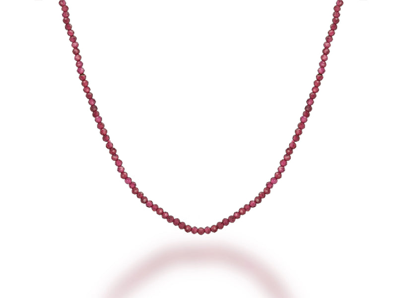 The Rachel Reid 28 Gemstone Bead Necklace features small, round, faceted red beads strung in a gentle curve on a white background, casting a soft shadow below.