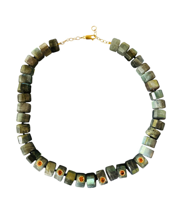 The Grand Labradorite Necklace by Jen Proudman features chunky square green labradorite stones, a gold clasp, and mandarin garnet-accented beads. This handmade piece forms a bold circular statement.