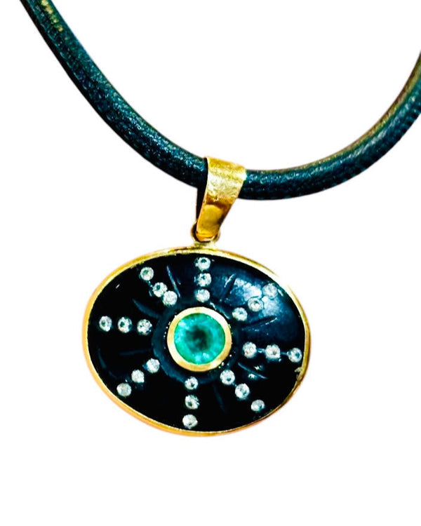 The SOL Pendant by Jen Proudman features an oval emerald center with diamond accents in a starburst on black onyx, set in a 14k gold frame and suspended from a dark cord necklace.