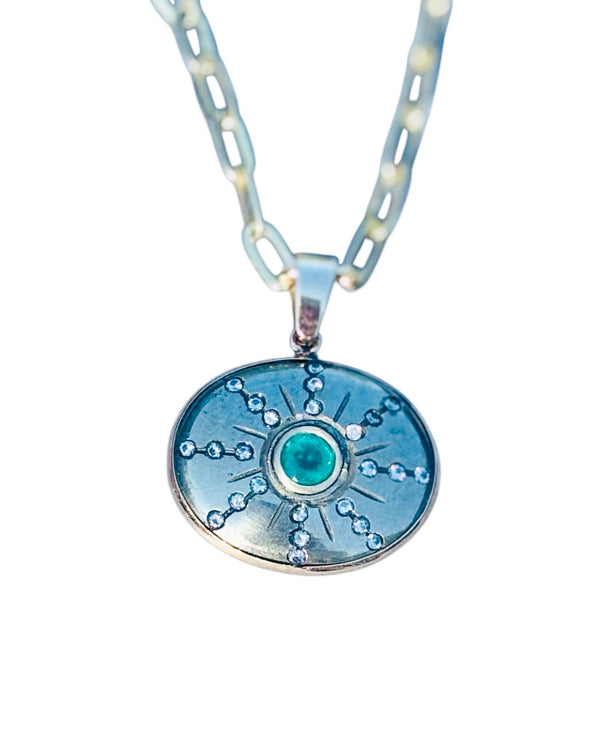 The Jen Proudman Sol Pendant features an oval silver design with a central green gemstone inspired by pyrite, emerald, and diamonds, radiating lines with clear stones, and a thick chain.