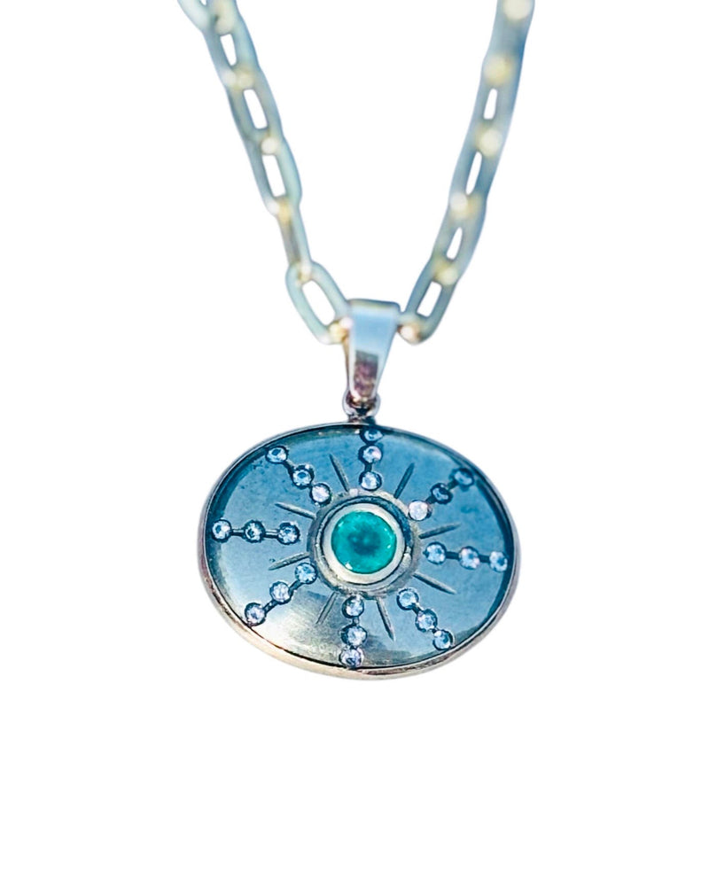 The Jen Proudman Sol Pendant features an oval silver design with a central green gemstone inspired by pyrite, emerald, and diamonds, radiating lines with clear stones, and a thick chain.