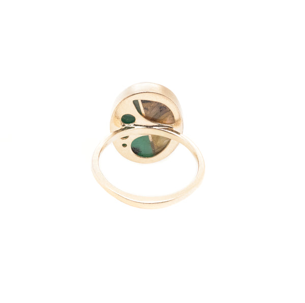 The Yin Yang Oval Ring by Karen Luvaas features a simple gold band and an oval bezel setting with a translucent stone, revealing green and brown hues visible from beneath.