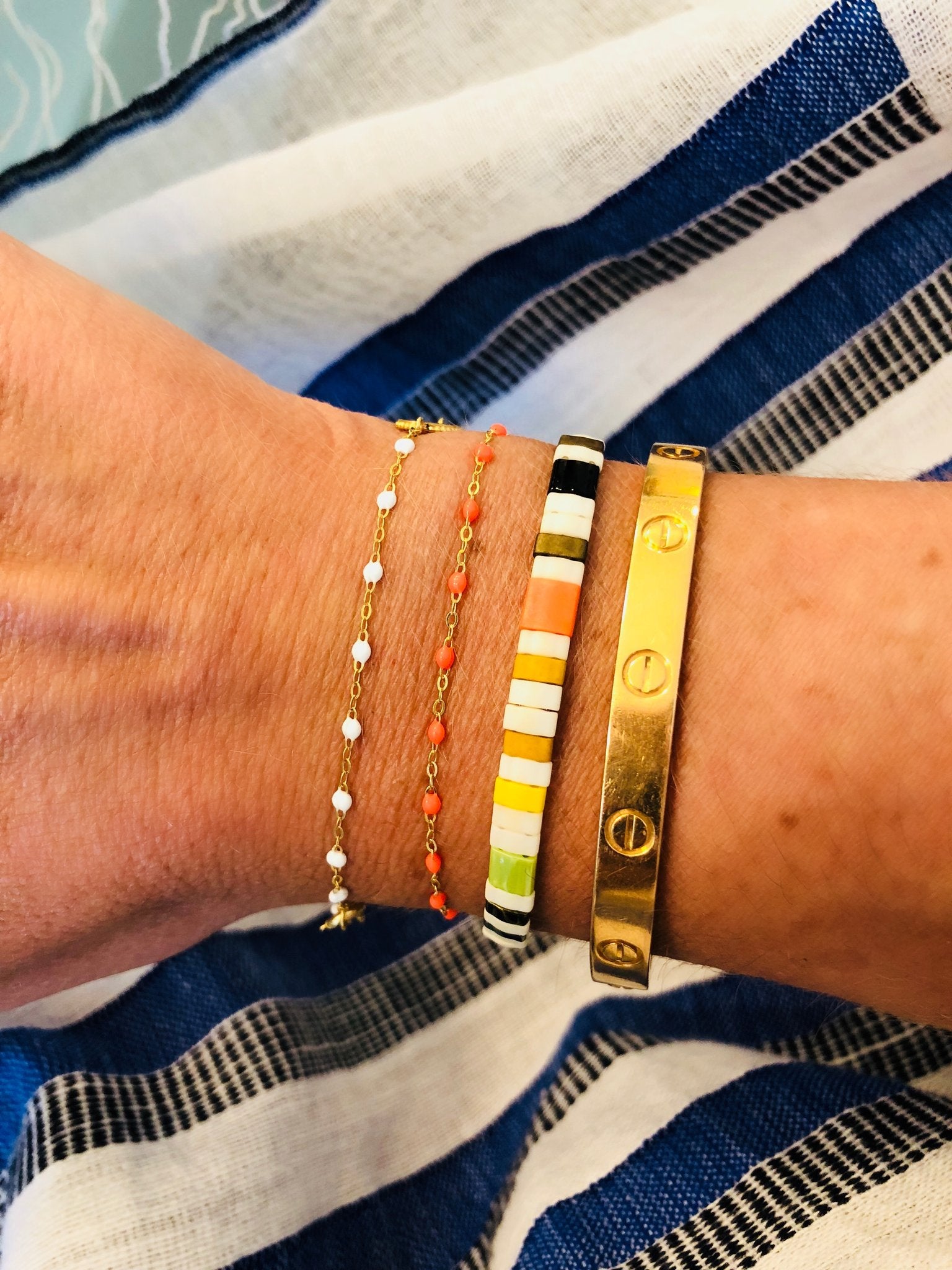 A wrist showcases a trio of elegant bracelets: a delicate Classic Gigi Bracelet by Gigi Clozeau, available in multiple colors; a vibrant multicolored beaded bracelet; and a wide gold bracelet featuring distinctive screw motifs. The backdrop features blue and white striped fabric, enhancing the luxury of these handmade pieces crafted in France for discerning jewelry enthusiasts.