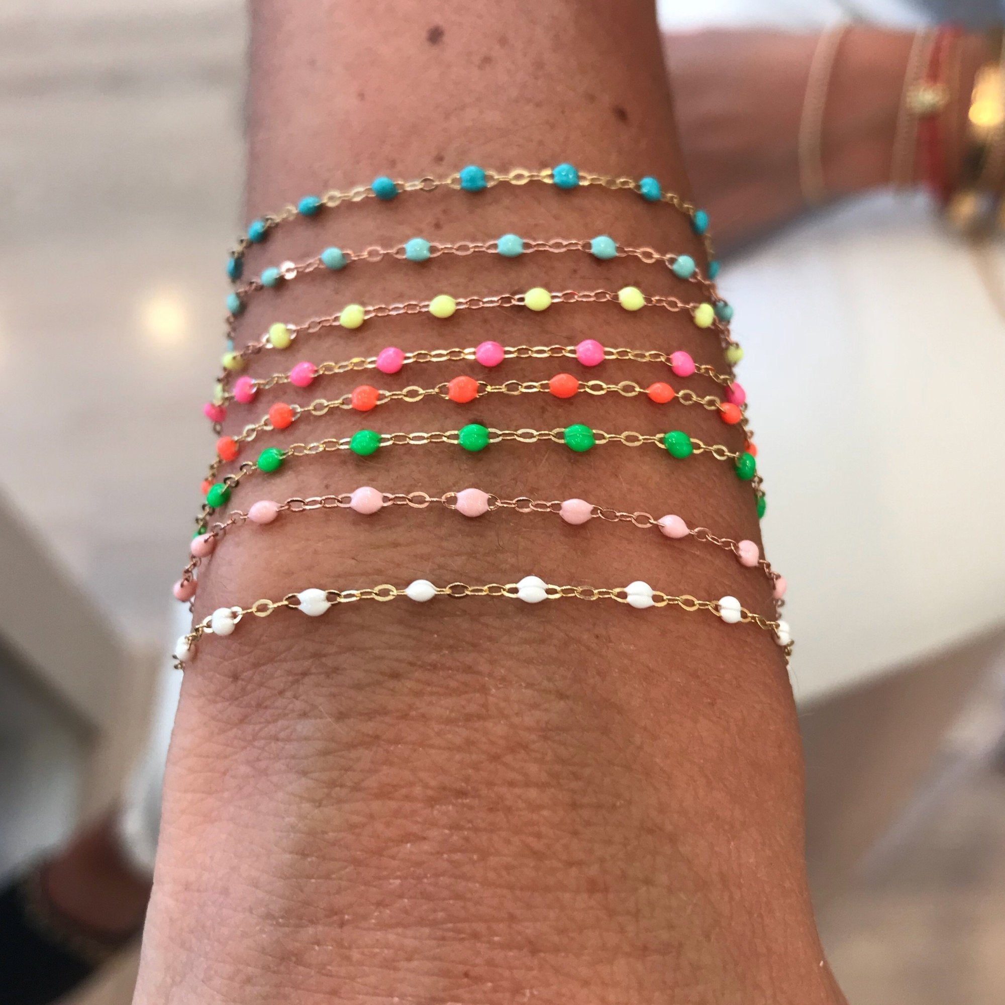 A wrist elegantly displays several colorful beaded bracelets attached to delicate gold chains. The adjustable Classic Gigi Bracelet by Gigi Clozeau, handcrafted in France from 18k gold, showcases an array of beads in blue, pink, red, green, yellow, and white hues for a vibrant and fashionable appearance.