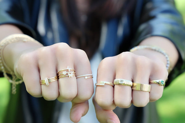 A person wearing a black jacket extends both fists, each adorned with multiple rings. Among them is the Rachel Reid Fluted Emerald Diamond Ring in 14K yellow gold, which adds an elegant touch against the blurred green background.